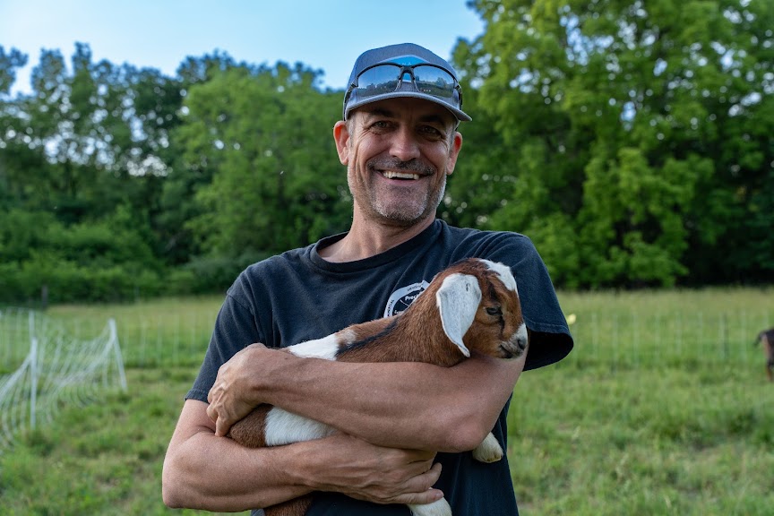 Sam smiling while holding a young goat outdoors